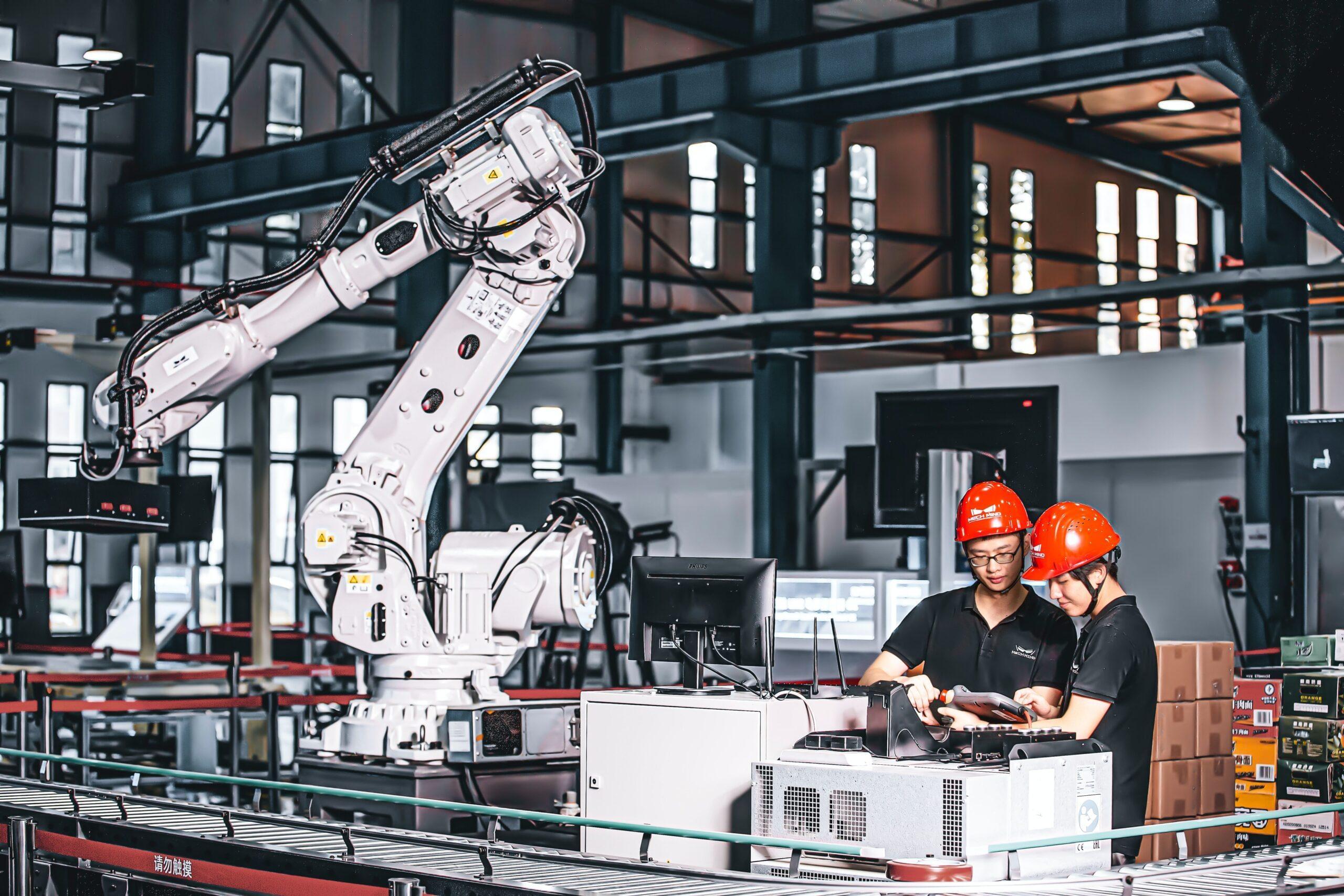 Two technicians wearing safety helmets operating and monitoring an industrial robotic arm in a modern factory setting.